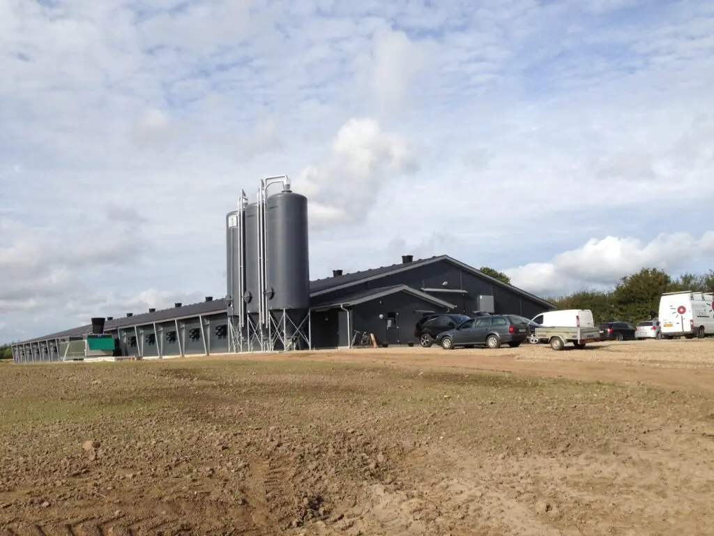 Modern dark gray poultry house with three large feed silos and parked vehicles on a farm site by LANDMECO.