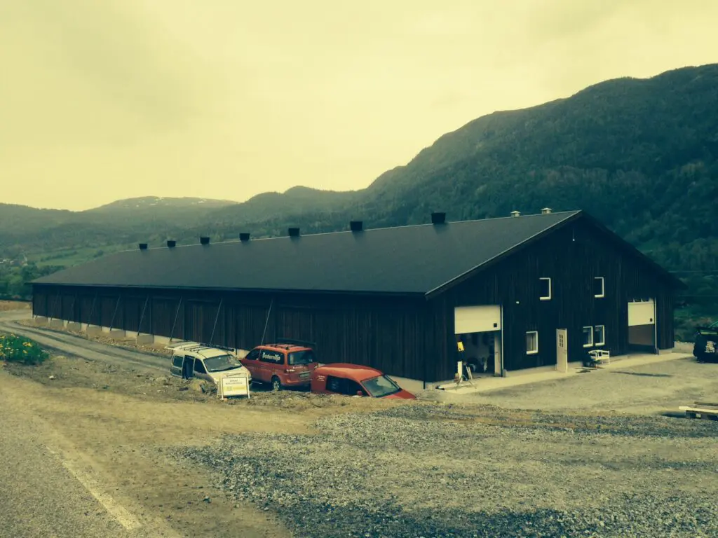Exterior view of a large dark poultry barn with service vehicles in a mountainous landscape.