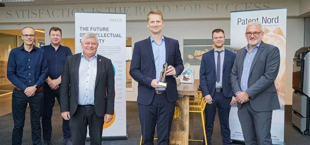 Six men in professional attire holding a trophy in front of Patent Nord and Zacco banners at LANDMECO headquarters.
