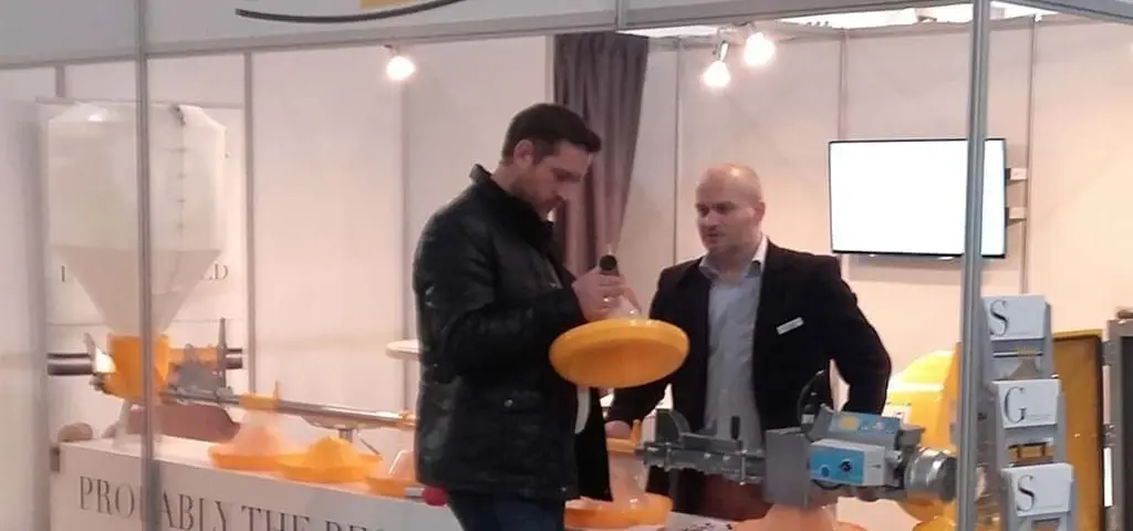 Professionals at LANDMECO Ferma Exhibition booth examining a yellow broiler pan feeding system.
