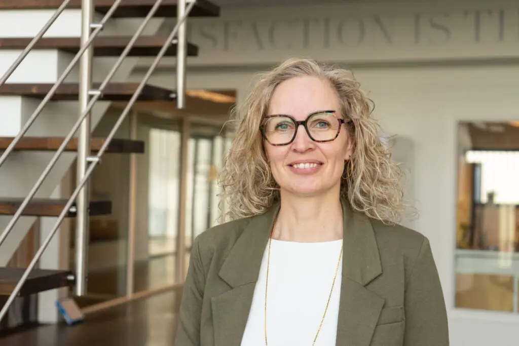 Henriette, Planning Specialist at LANDMECO, wearing glasses and a green blazer in a modern office setting.