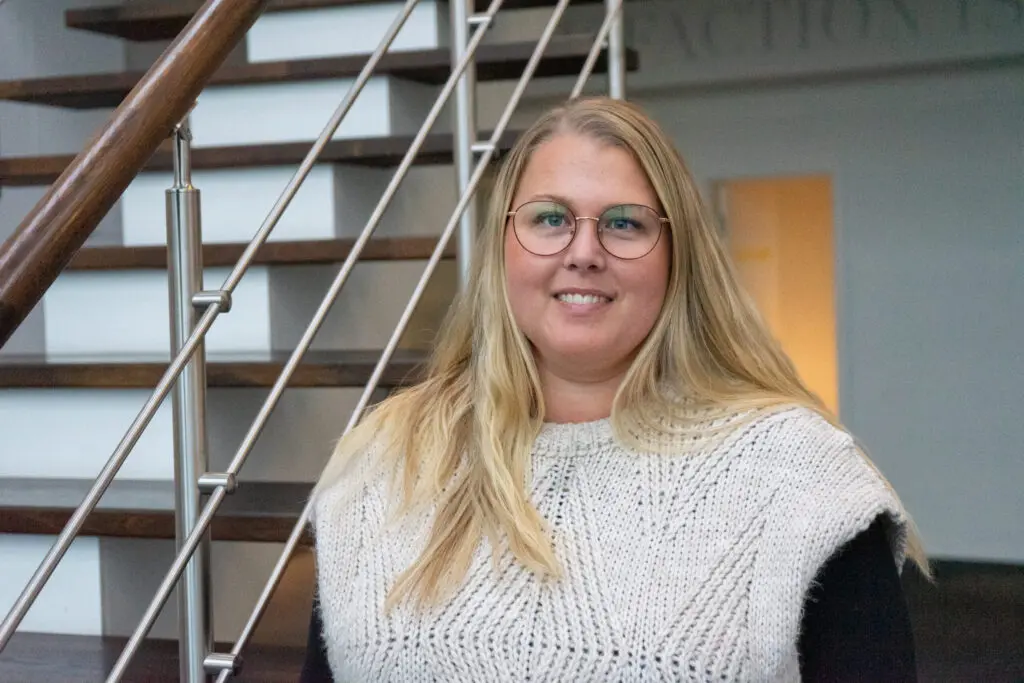 Line Korsholm, PA to Production Manager at LANDMECO, smiling in front of an office staircase in Ølgod.