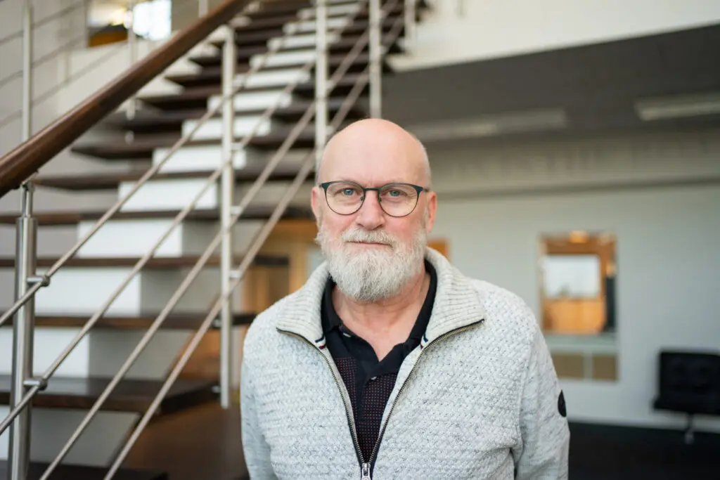 Svend, LANDMECO Technical Support team member with a white beard and glasses, standing in a modern office near stairs.