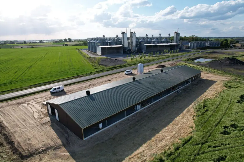 Aerial drone view of a modern poultry house and grain silos in a green field landscape.