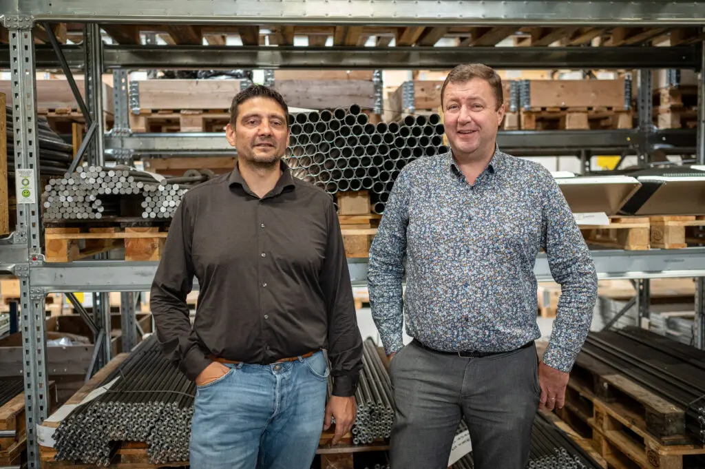 Repræsentanter fra Landmeco og PG Industri foran reoler med metalrør og drejede komponenter på lageret.