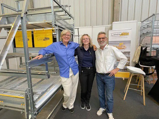 Three LANDMECO representatives standing in front of galvanized steel poultry housing equipment and brand banners at a trade show.