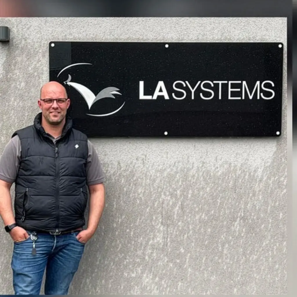 John Buck, Managing Director of LA Systems, standing in front of the LA Systems company sign on a grey wall.