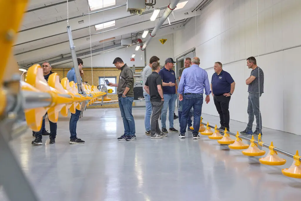 A group of men standing in a large production facility, looking at a row of yellow poultry feeders during a dealer seminar.