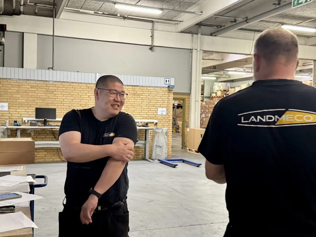 To lagerarbejdere i sorte LANDMECO t-shirts taler sammen i et lyst lagermiljø.