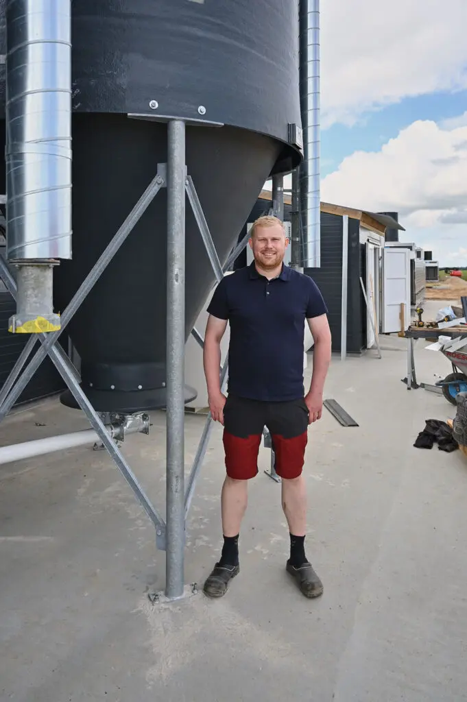 Mathias Højgaard står foran en stor sort fodersilo ved en moderne kyllingestald under opførelse.