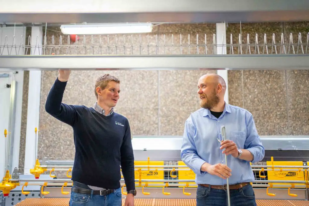 Two men in a professional setting discussing poultry rearing equipment, with LANDMECO systems in the background.