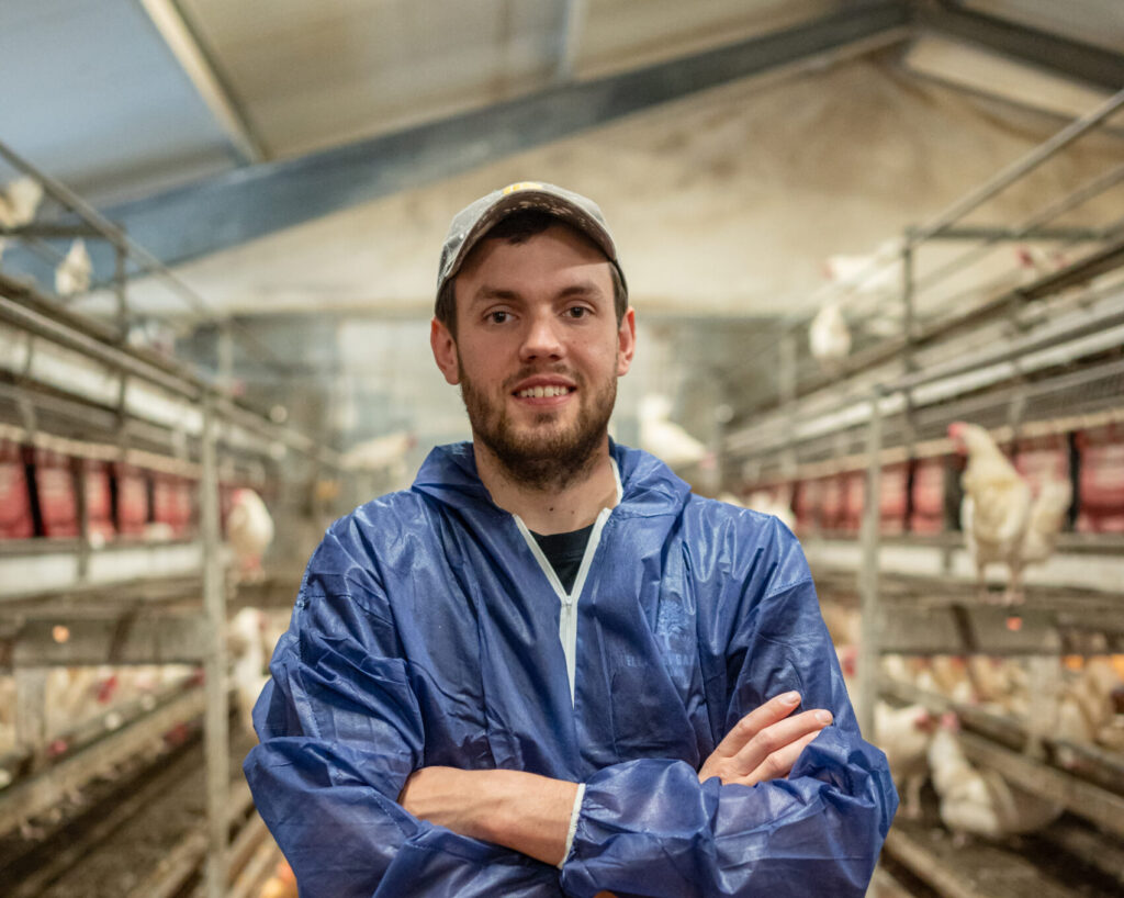 Danish farmer in a blue suit standing in a LANDMECO layer aviary system house with tiered poultry equipment.
