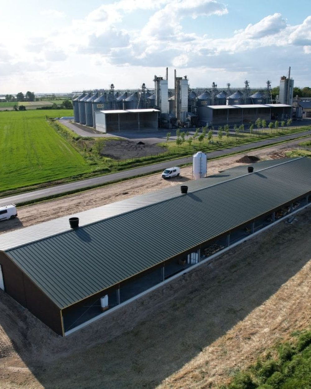 Aerial drone view of a modern poultry house and grain silos in a green field landscape.