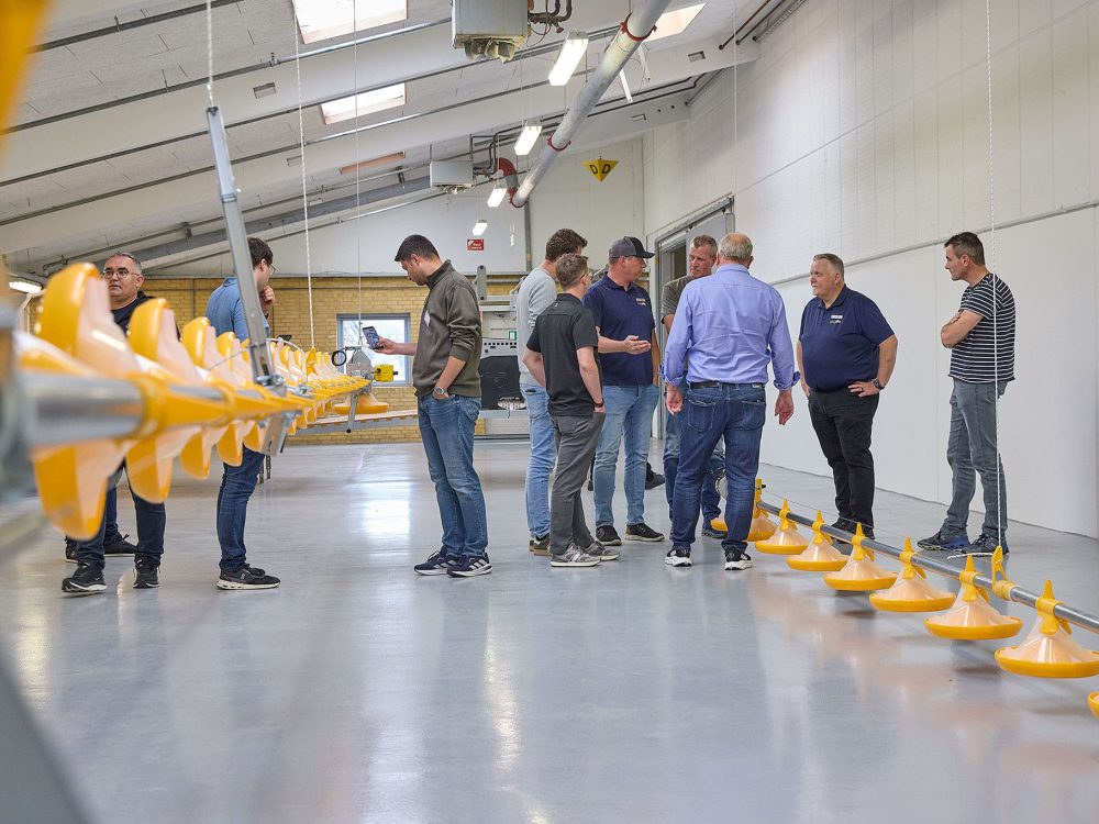 A group of men standing in a large production facility, looking at a row of yellow poultry feeders during a dealer seminar.