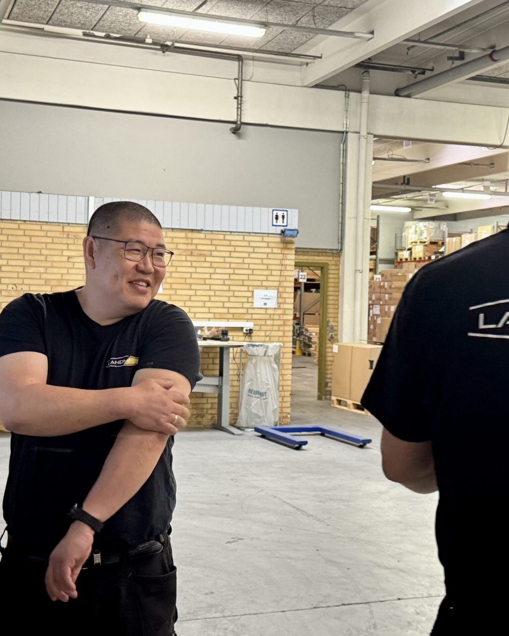 To lagerarbejdere i sorte LANDMECO t-shirts taler sammen i et lyst lagermiljø.