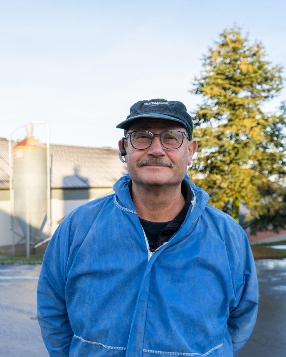 Poultry producer in blue overalls and LANDMECO cap standing before a farm with silos and production houses.