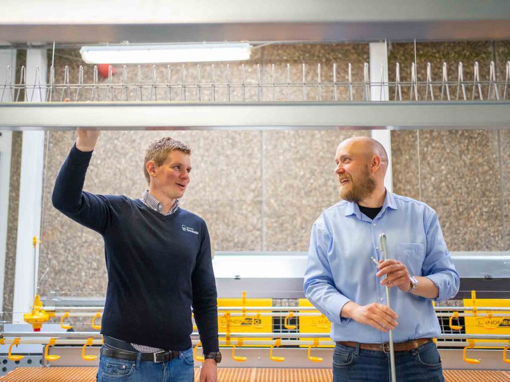 Two men in a professional setting discussing poultry rearing equipment, with LANDMECO systems in the background.