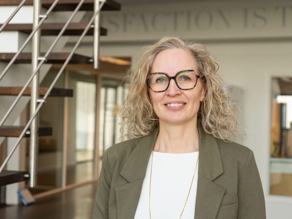 Henriette, Planning Specialist at LANDMECO, wearing glasses and a green blazer in a modern office setting.