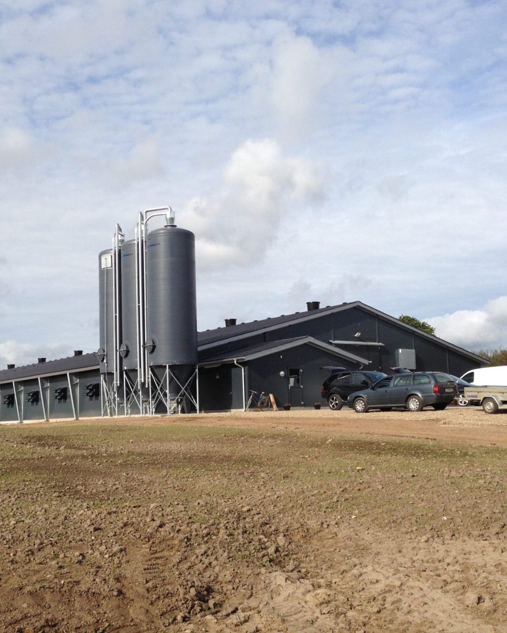 Modern dark gray poultry house with three large feed silos and parked vehicles on a farm site by LANDMECO.