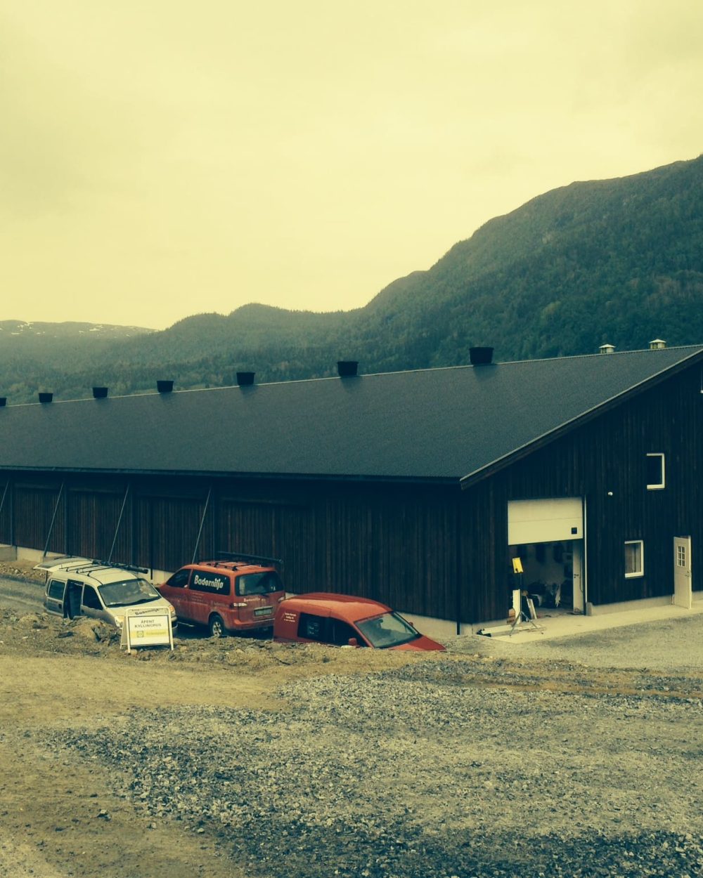Exterior view of a large dark poultry barn with service vehicles in a mountainous landscape.
