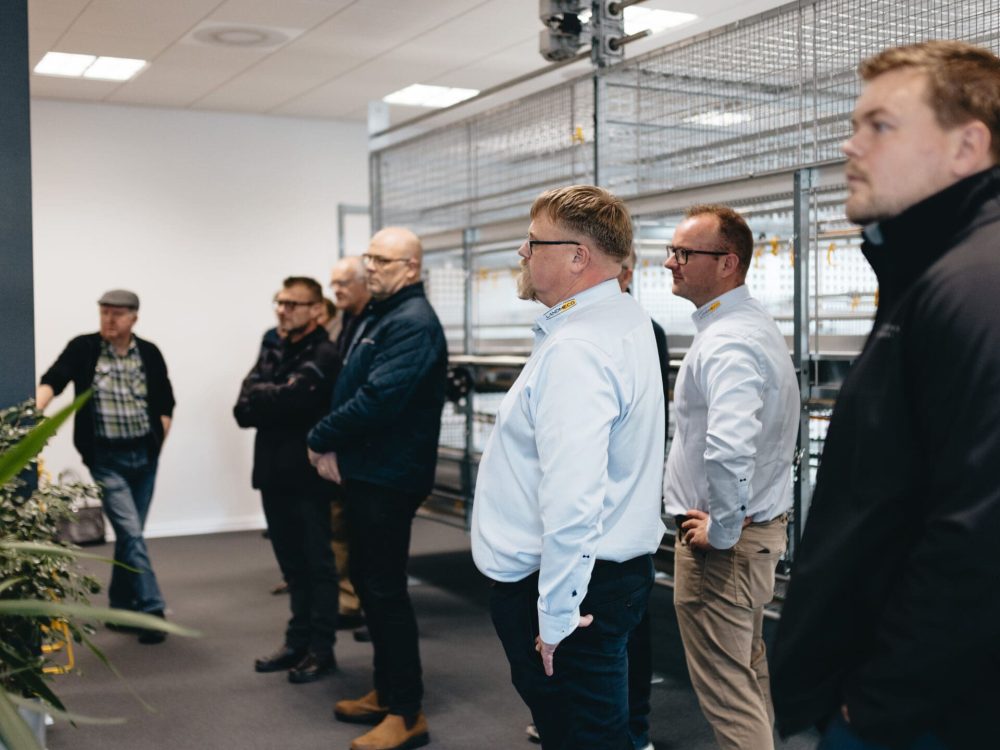 En gruppe mænd i et showroom kigger på LANDMECOs moderne staldsystemer til fjerkræ under Fjerkrædagen 2025.