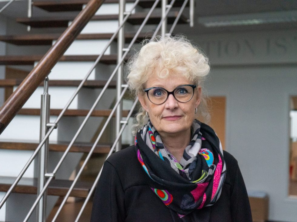 Inge Nørgaard from LANDMECO, wearing glasses and a colorful scarf, standing by stairs in the Ølgod office.