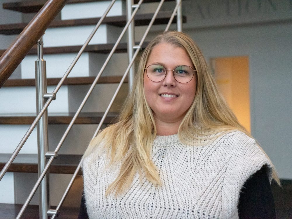 Line Korsholm, PA to Production Manager at LANDMECO, smiling in front of an office staircase in Ølgod.