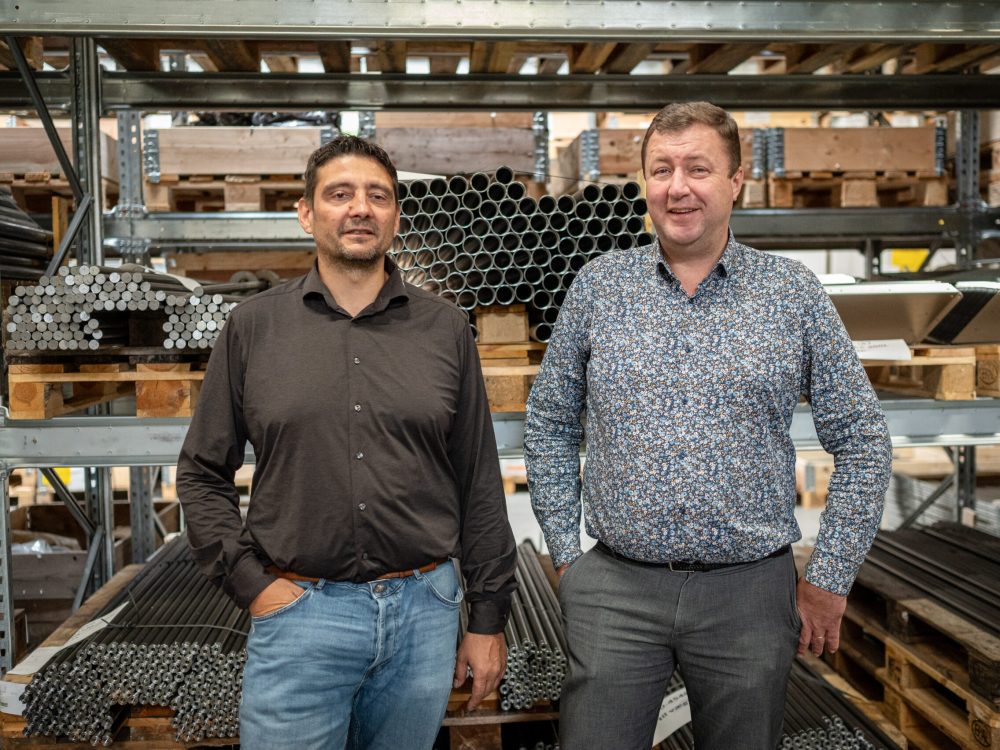 Repræsentanter fra Landmeco og PG Industri foran reoler med metalrør og drejede komponenter på lageret.