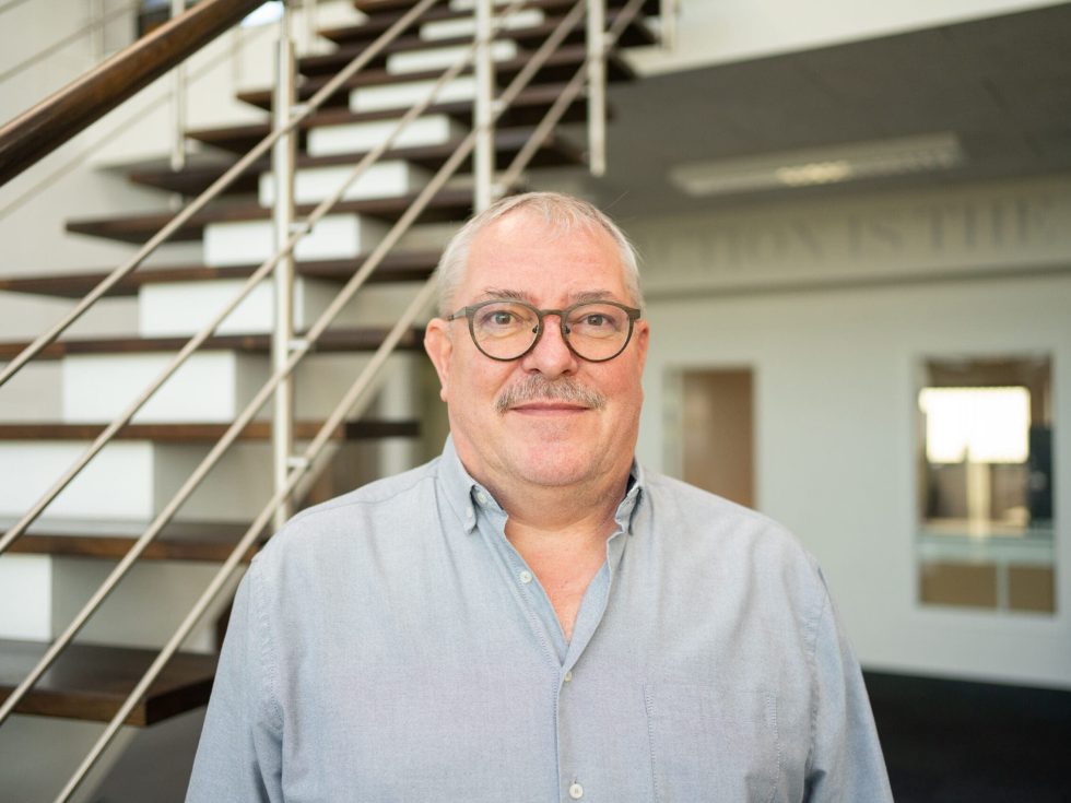 Peter Buhl of LANDMECO, wearing glasses and a blue shirt, standing in a modern office with a wooden staircase.