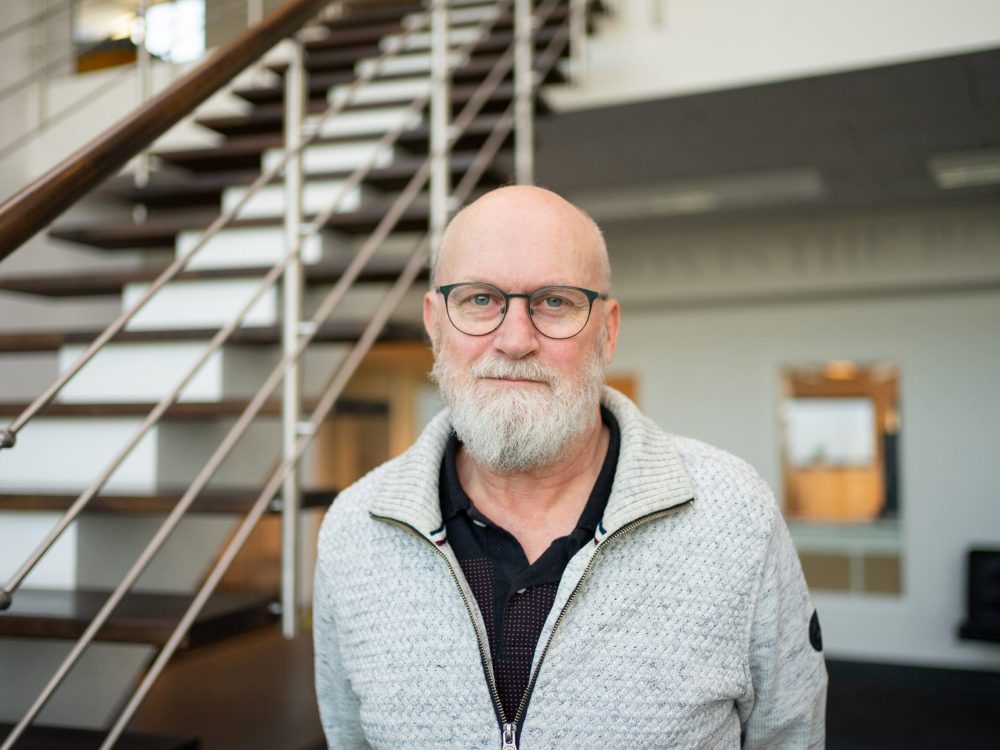 Svend, LANDMECO Technical Support team member with a white beard and glasses, standing in a modern office near stairs.
