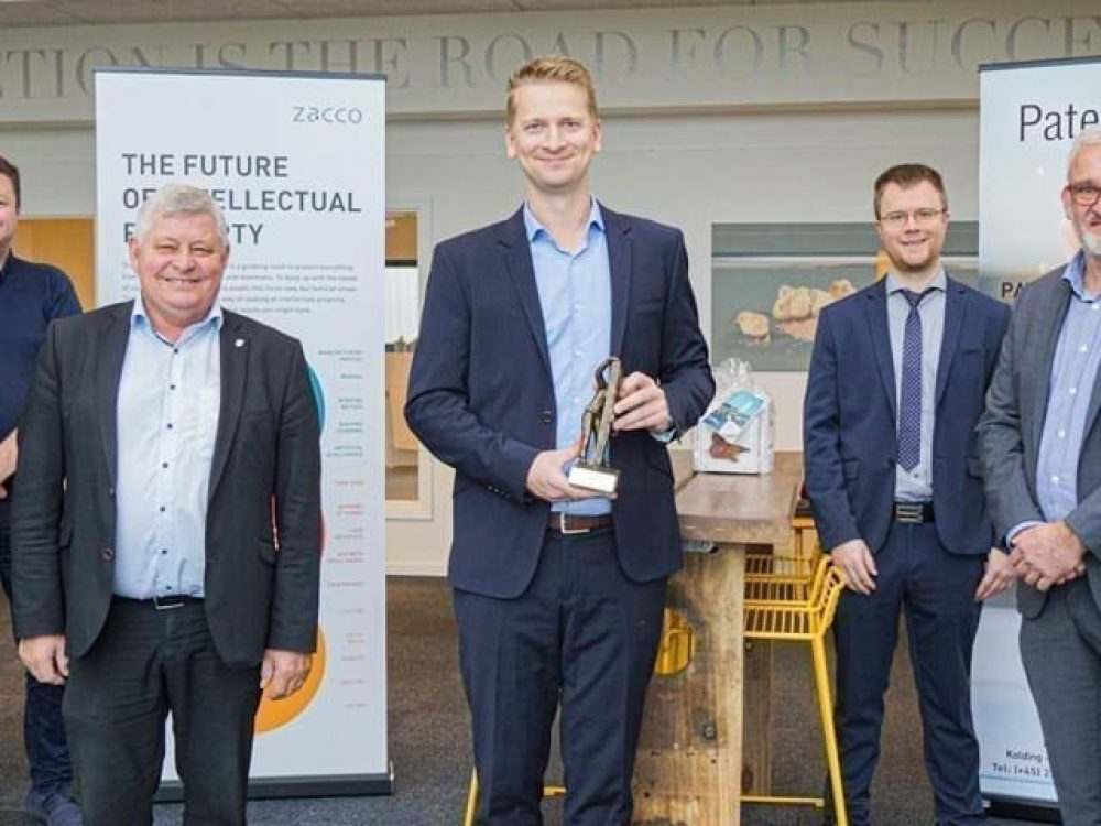 Six men in professional attire holding a trophy in front of Patent Nord and Zacco banners at LANDMECO headquarters.