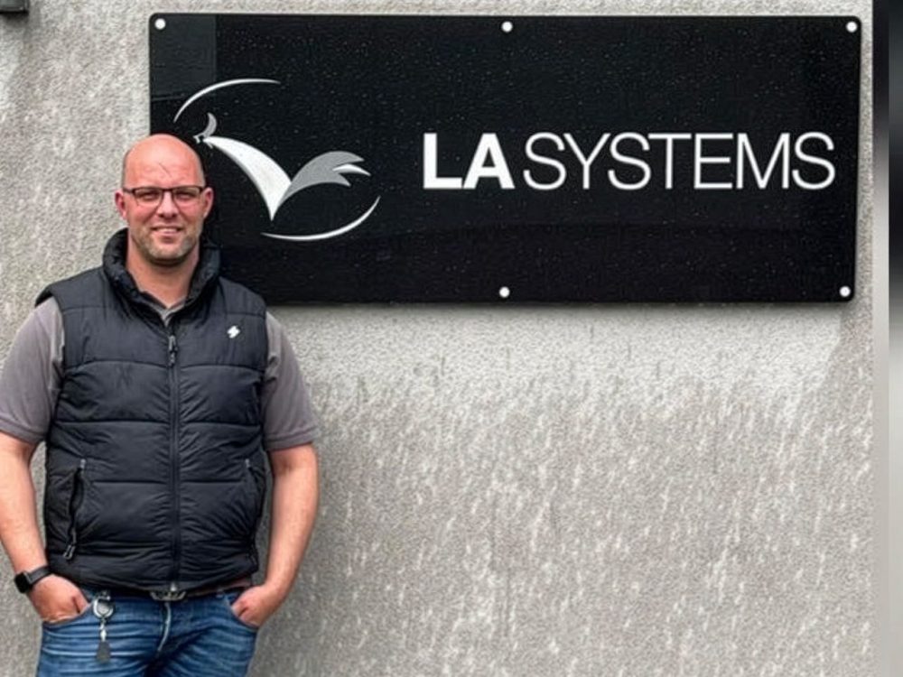 John Buck, Managing Director of LA Systems, standing in front of the LA Systems company sign on a grey wall.