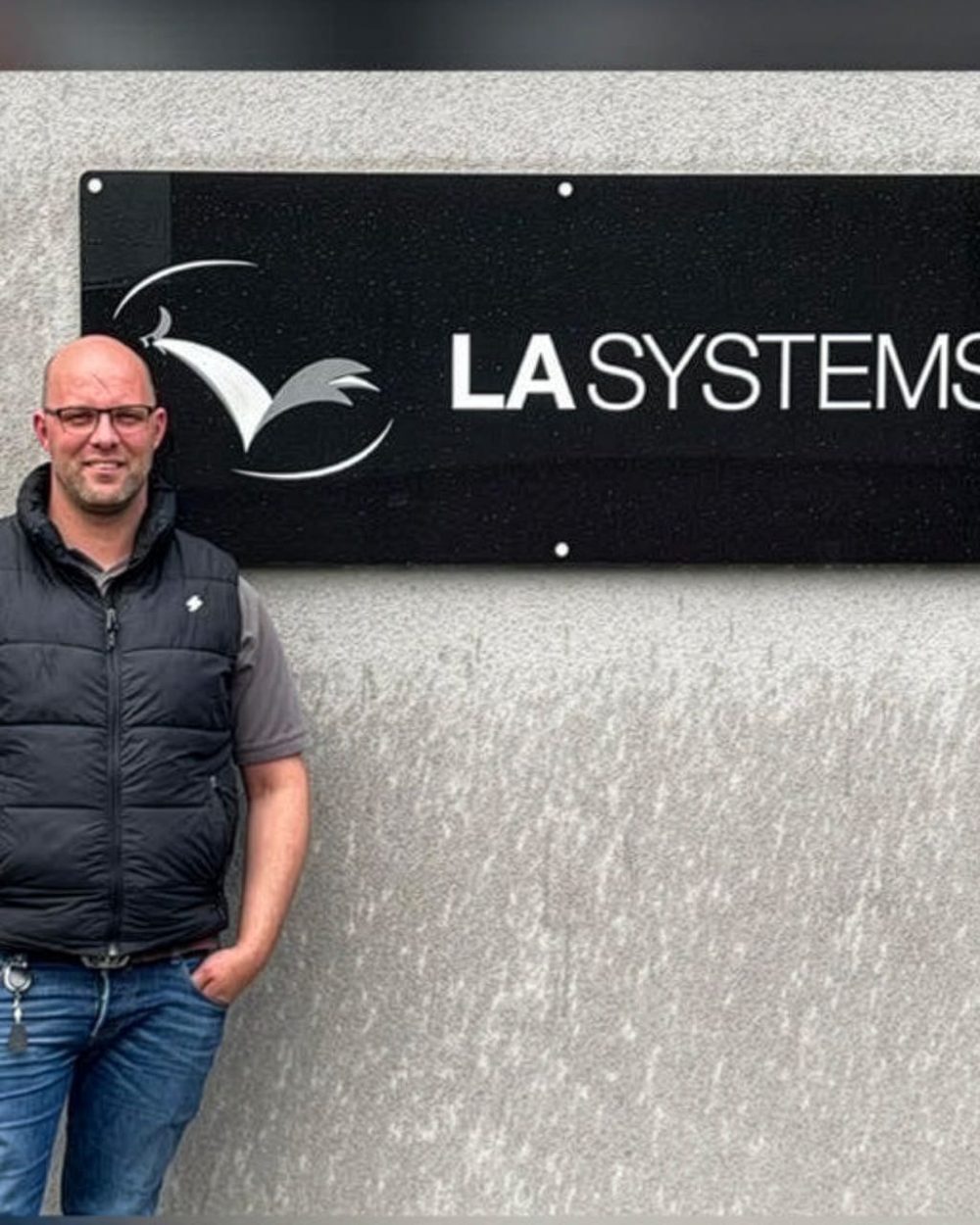 John Buck, Managing Director of LA Systems, standing in front of the LA Systems company sign on a grey wall.