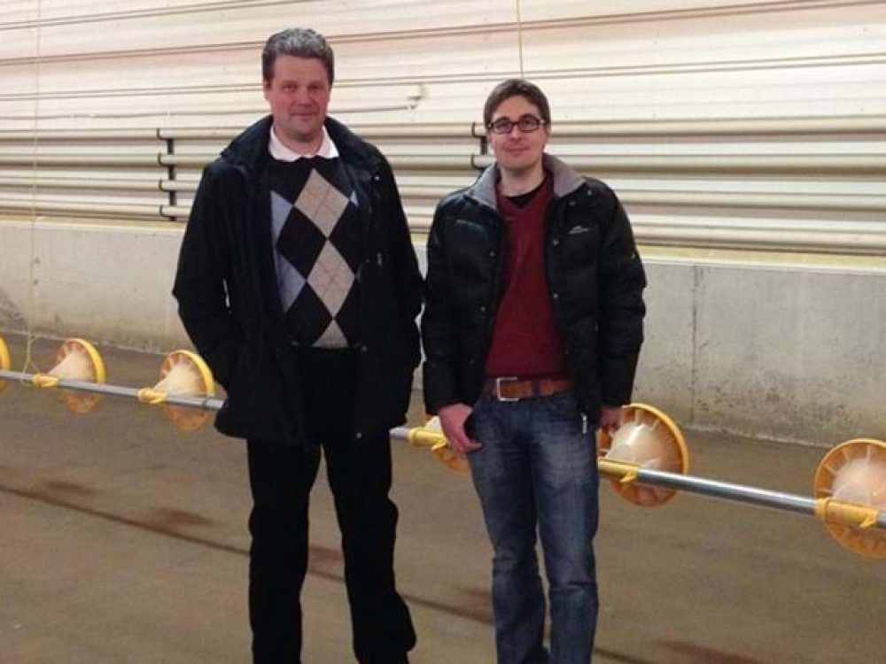 Two experts standing next to a LANDMECO automated broiler pan feeding system in a modern poultry house.